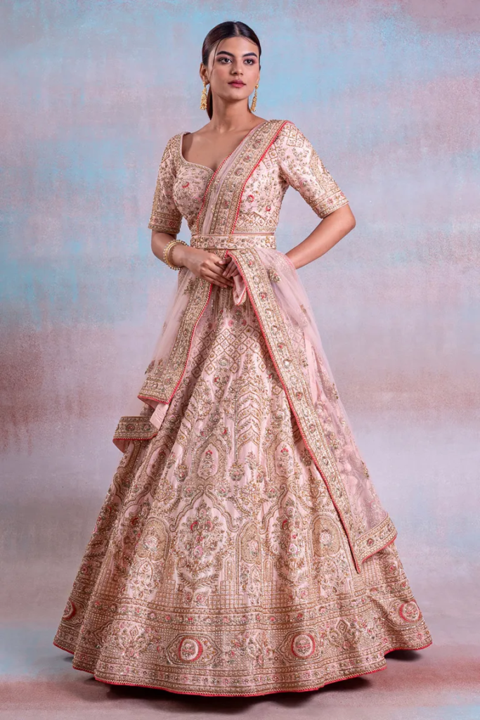 This exquisite pink silk lehenga choli is a masterpiece of embroidery and sequins work. The pale pink flared lehenga, with a length of 112 cm, features intricate Cutdana, Zari, Sequins, Pearl, and Resham work, exuding elegance and purity. The net dupatta in a matching pale pink shade is adorned with a gold border and the Cutdana, Zari, Sequins, Pearl & Resham Work, adding grace to the ensemble. The readymade choli, also in pale pink silk, showcases a tie-back neck and leaf neck pattern, with elbow sleeves, and is embellished with the same Cutdana, Zari, Sequins, Pearl & Resham Work. This ensemble is a perfect choice for a grand occasion, radiating charm and sophistication. Complete the look by wearing Diamond jewelry and bangles. There might be little color variations in the image and original product due to photographic lightings or your monitor settings.