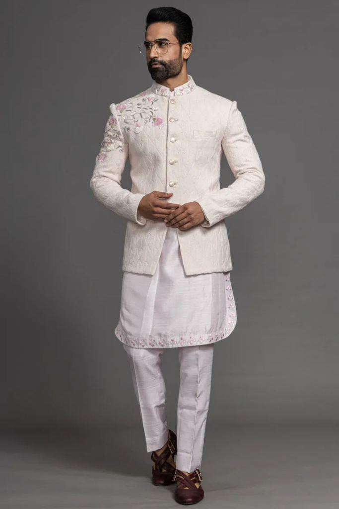 This exquisite party wear suit ensemble is designed to make a bold and fashionable statement at special occasions. The slim-fit suit, tailored from luxurious jute fabric in a unique creamy pink color, features a stand-up collar and long sleeves, exuding a contemporary and sophisticated charm. The suit is beautifully adorned with woven, bullion, and Zardosi work, creating a rich and intricate pattern that adds a touch of opulence and artistry to the overall look. Paired with straight-cut cotton silk trousers in white, it creates a harmonious balance between style and comfort. The ensemble is designed for formal events and gatherings, showcasing a blend of tradition and modern aesthetics. The kurta, crafted from cotton silk, complements the suit perfectly with its Resham work, offering a cohesive and elegant appearance. This ensemble is a perfect choice for parties and formal gatherings where you want to make a memorable and stylish impression, showcasing a blend of tradition and modern aesthetics while maintaining a refined and opulent charm. There might be little color variations in the image and original product due to photographic lightings or your monitor settings.